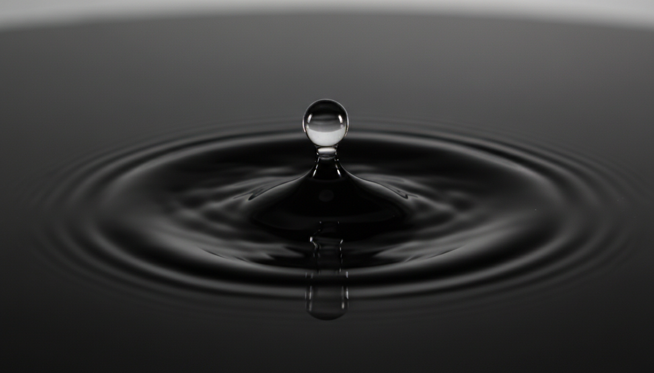 A macro photograph of a single water droplet hitting a perfectly smooth black glass surface, creating one clean concentric ripple.