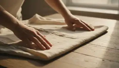 Close-up, photorealistic shot of hands gently smoothing out the wrinkles of a soft, light-colored linen shirt on a wooden table. Warm, golden hour lighting highlighting the intricate texture of the fabric. Peaceful atmosphere, high detail on the fabric weave, conveying tactile grounding and calmness.