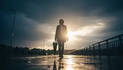 Fotografía íntima y cálida. Una persona caminando sola por una acera mojada justo después de que ha terminado una gran tormenta. Ha cerrado su paraguas y mira hacia el cielo donde las nubes oscuras se están abriendo para dejar pasar unos rayos de sol cálido y brillante. Su rostro muestra una sonrisa sincera de alivio y paz interior. Reflejos de luz en los charcos del suelo. Estilo cinematográfico inspirador.