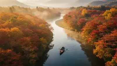 Una toma aérea cenital espectacular y fotorrealista de un pequeño bote de madera flotando suavemente a la deriva por un río serpenteante. El río está rodeado de árboles de arce japonés en tonos rojos y dorados otoñales, con una ligera niebla. Una persona está sentada en el bote, relajada, sin remar, simplemente dejándose llevar por la corriente con total confianza. Hora dorada, calidad National Geographic.