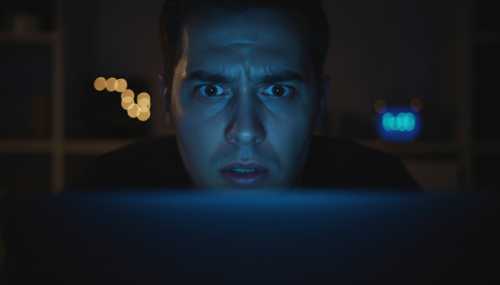 A photorealistic image of a person staring at a glowing laptop screen late at night, their face illuminated by the harsh blue light, showing subtle signs of panic and hypervigilance, representing the modern 'fight or flight' response. Cinematic lighting, shallow depth of field, dramatic shadows.