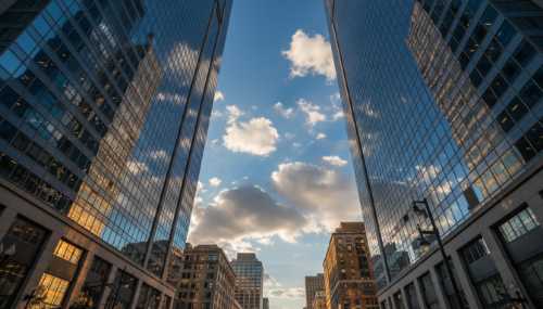 A low-angle photorealistic shot from a city street looking straight up. Tall glass skyscrapers frame a vibrant deep blue sky filled with fluffy white clouds. The golden sunlight of late afternoon reflects off the buildings. A sense of awe, peace, and expansiveness. High resolution, vivid and bright colors.
