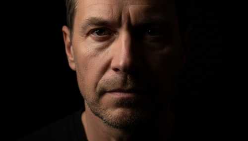 Retrato fotorrealista con técnica de claroscuro (chiaroscuro): Un hombre mirando fijamente hacia la cámara con una expresión de valentía y aceptación. La mitad de su rostro está bañada por una luz dura y cálida, mientras que la otra mitad se funde gradualmente con una oscuridad densa e infinita. Gran detalle en los poros de la piel, concepto de trabajo de sombra psicológico.