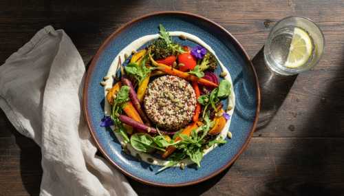 Top-down view of a vibrant, beautifully plated plant-based meal on a rustic ceramic plate. The dish features colorful roasted vegetables, fresh greens, quinoa, and a creamy vegan dressing. Set on a wooden dining table with a linen napkin and a glass of water. Food photography, highly appetizing, natural lighting, sharp and photorealistic.