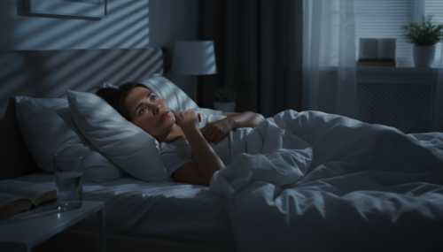 Photorealistic image of a woman lying in bed at night in a dark room, illuminated only by moonlight coming through the blinds. Her eyes are wide open, staring straight up at the ceiling, visibly anxious and unable to sleep due to racing thoughts. Cinematic lighting, dramatic shadows, highly detailed and realistic bedroom environment.