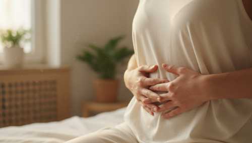 Fotografía fotorrealista e íntima. Un primer plano de las manos de una persona descansando suavemente sobre su abdomen bajo, justo debajo del ombligo. Lleva ropa cómoda y suave de algodón en tonos neutros. La iluminación es suave y cálida, sugiriendo un momento de calma, respiración profunda y conexión mente-cuerpo en una habitación serena.