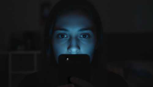 High-quality photograph of a person in a dark room illuminated only by the cold blue light of a smartphone screen. The person's face shows emotional numbness and detachment. Cinematic realism, 85mm lens, sharp focus on the face highlighting the concept of emotional avoidance.