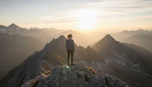 A photorealistic image of a person standing on a mountain peak at sunrise, looking ahead with a confident, calm, and grounded expression. The lighting is warm and golden, symbolizing realistic optimism, mental resilience, and the biological translation of a welcoming environment. Wide angle, beautiful natural scenery.