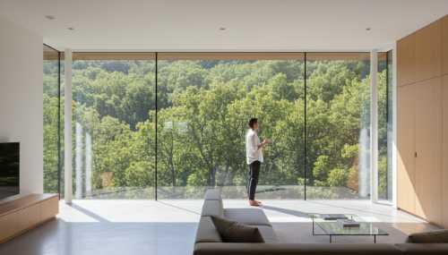 A bright, sunlit modern room with large floor-to-ceiling windows. A person is standing by the glass, hands relaxed, looking out at a vibrant green forest, completely at ease and grounded in the present moment. The atmosphere feels liberating, peaceful, and clear. Crisp photography, realistic textures, architectural digest style.