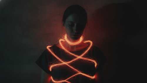 A conceptual, highly emotional photographic portrait of a person in shadow, with abstract red and orange light fibers wrapping around their spine and chest, symbolizing the physical manifestation of emotional stress and chronic pain. The person has a serene but intense expression, suggesting deep introspection. Moody, cinematic lighting, dramatic shadows, realistic textures, 8k resolution.