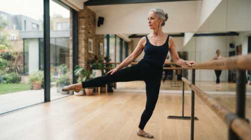 An active, graceful older woman in her 60s practicing ballet at a wooden barre. She has a serene, sharp expression of deep concentration, wearing simple athletic wear in a modern, brightly-lit wellness studio. Realistic photography, natural skin texture, inspiring and healthy lifestyle.