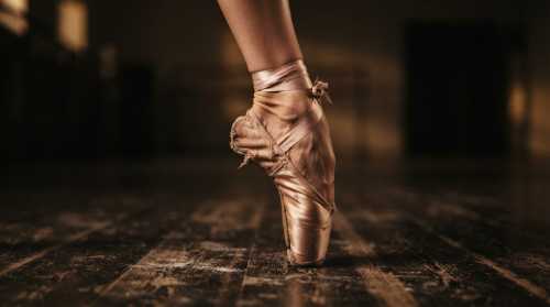 Extreme macro close-up of a ballet dancer's foot en pointe on a dark wooden studio floor. The satin pointe shoe shows fine texture and slight wear. The background is softly blurred with warm, moody lighting focusing entirely on the extreme balance, tension, and micro-adjustments of the foot.
