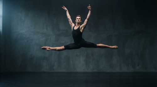 A powerful male ballet dancer captured in high-speed action photography mid-air in a perfectly symmetrical grand jeté. His muscles are defined, showing peak athletic form. Clean, minimalist studio background with cool dramatic side-lighting highlighting the anatomical symmetry.