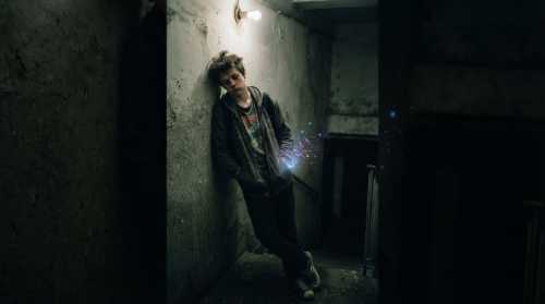 A moody, realistic portrait of an exhausted teenager leaning against a cold concrete wall, looking downcast. Overhead, a faint, dim lightbulb flickers, while digital sparks faintly emanate from a phone screen in their pocket, representing the desperate search for dopamine. Desaturated colors, gritty texture, expressing clinical anhedonia.